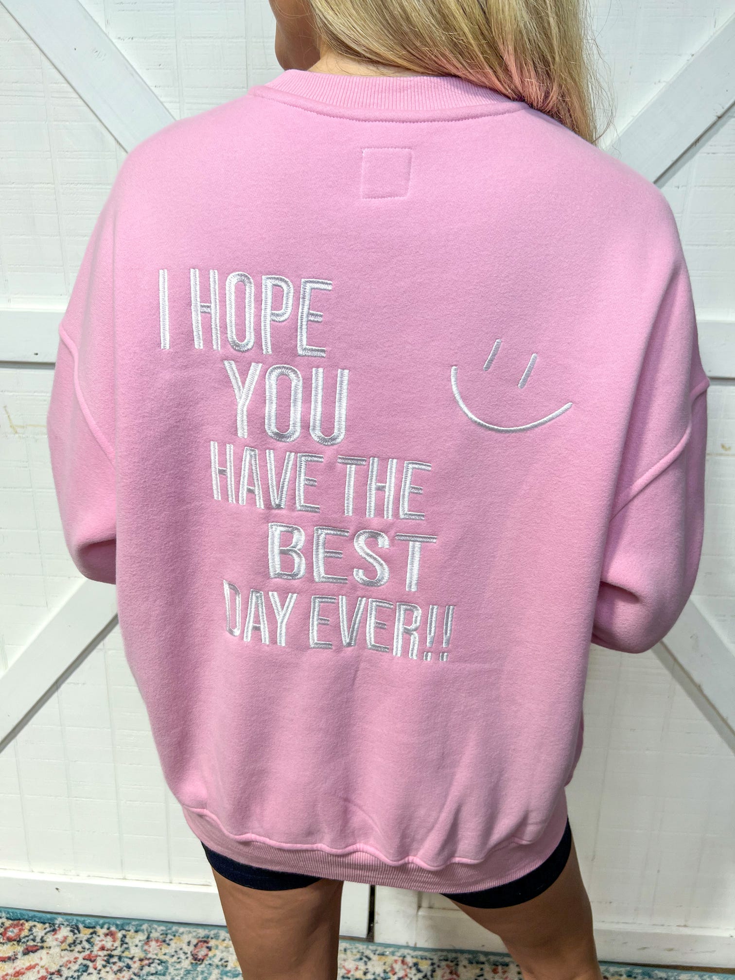 Up close photo of the back of the pink sweatshirt embroidered saying “I hope you have the best day ever” on the back along with another smiley face 