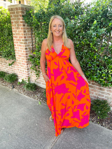 A woman modeling our orange maxi dress with magenta flowers all over. The dress has a v-neck and ties around the neck.