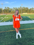 Woman modeling an orange sleeveless v-neck short dress with a white bow at the center of the v-neck. She's standing on a football field.