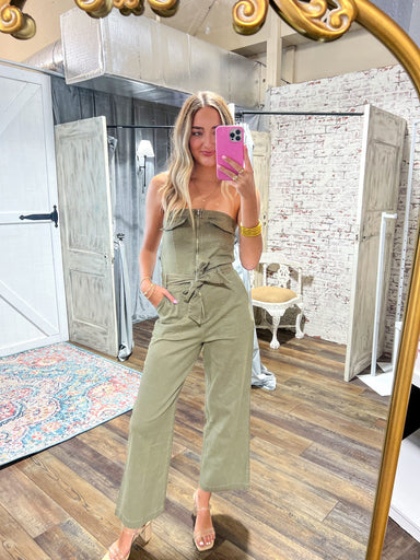 Mirror photo of a woman modeling our olive green strapless jumpsuit that zips and has a tie bow at the waist and wide leg pants.