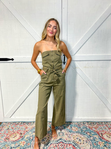 Woman modeling our olive green strapless jumpsuit that zips and has a tie bow at the waist and wide leg pants.