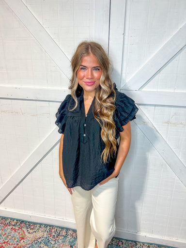 Photo of a woman wearing our black v-neck, short sleeve blouse with ruffles on the sleeves. She's wearing it with our Maddi cream Jeans. 