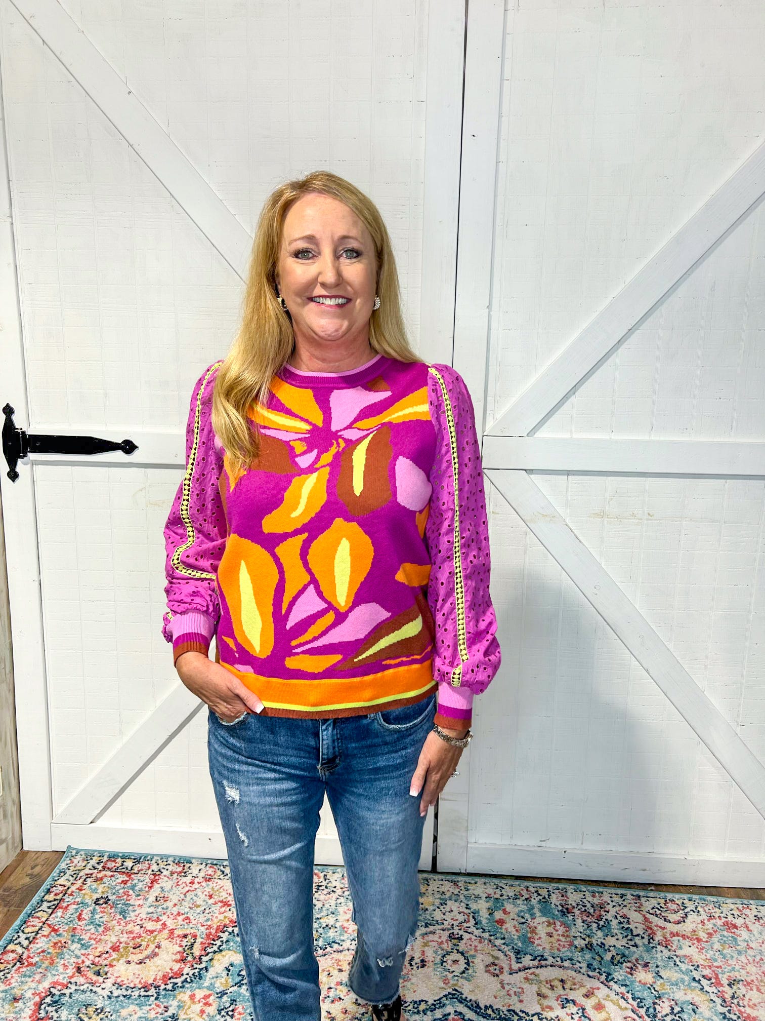 Photo of a mature woman modeling a purple thin sweater with eyelet lace sleeves. It has a floral design in lavender, orange and yellow.