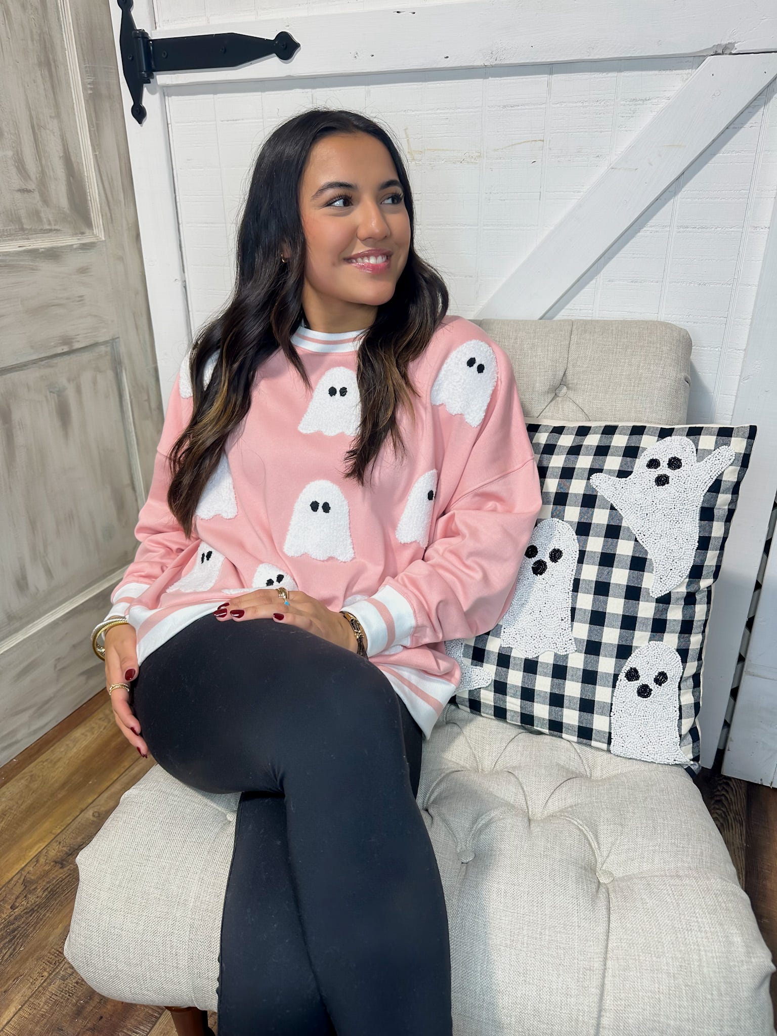 Young woman sitting on a chair modeling our light pink sweatshirt that is covered in white sequin ghosts. The chair also has a black and white checkered square pillow with white sequin ghosts.