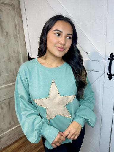 Up close photo our aqua blue long sleeve top featuring a suede taupe star with brass buttons around the trim of the star big on the front. 