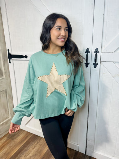 Young woman modeling our aqua blue long sleeve top featuring a suede taupe star with brass buttons around the trim of the star big on the front. 