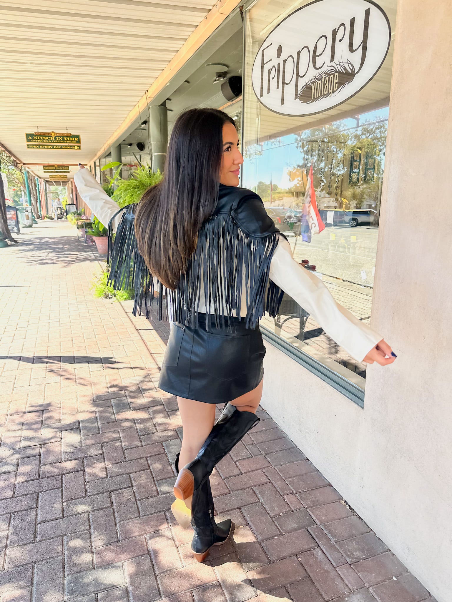 A woman is modeling the back of our cream faux leather jacket featuring long black fringe at the top of the front and back