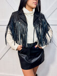 A woman is modeling our cream faux leather jacket featuring long black fringe at the top of the front and back