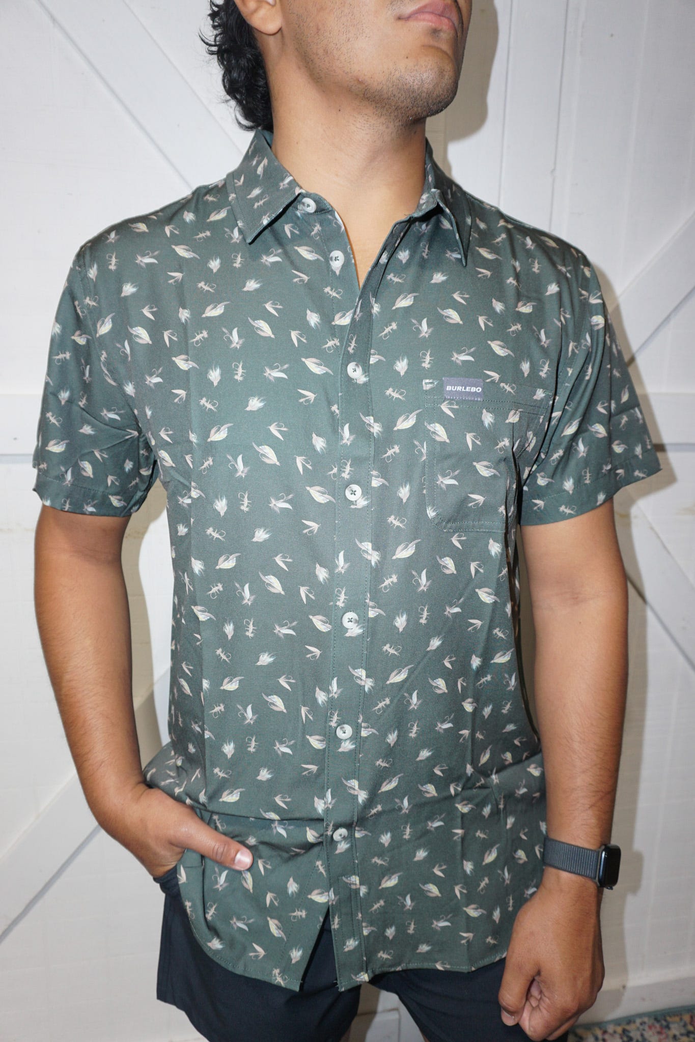 An up close photo of our dark green performance button up short sleeve shirt with several different fishing lures all over the shirt.