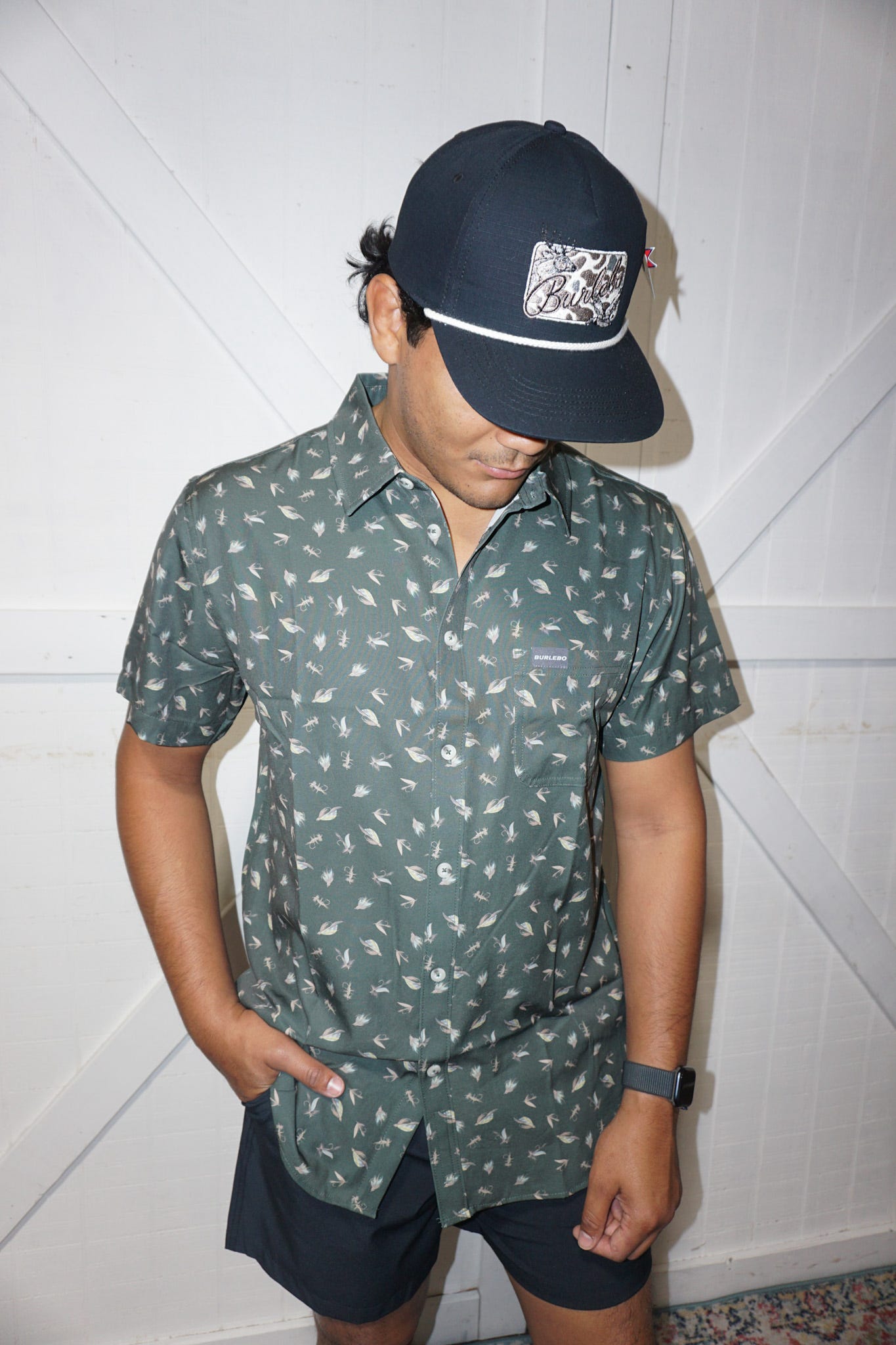 A male modeling our dark green performance button up short sleeve shirt with several different fishing lures all over the shirt.