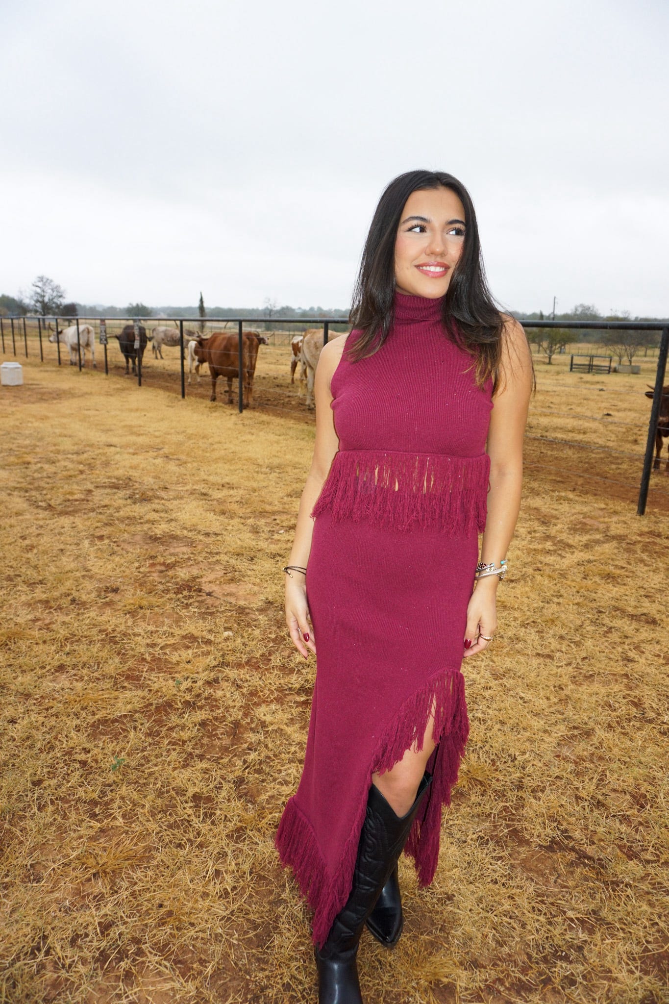 Another view of our matching two piece top and skirt set in a burgundy color. Made of sweater material, the top is sleeveless with a mock neck and fringe on the bottom. The long skirt is of the same material with fringe on the bottom and a slit on one side. The model is wearing it with black cowboy boots and is standing in a field with cows in the background.