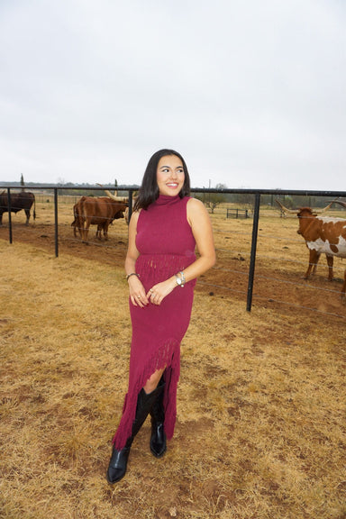 Woman modeling our matching two piece top and skirt set in a burgundy color. Made of sweater material, the top is sleeveless with a mock neck and fringe on the bottom. The long skirt is of the same material with fringe on the bottom and a slit on one side. The model is wearing it with black cowboy boots and is standing in a field with cows in the background.