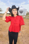 A woman posing in a field with a silo in the background modeling an up close photo of our scarlet red blouse that ties down the middle with bows and has bubble sleeves. She is wearing the top with a black denim long skirt with a front slit, a black cowboy hat and boots. 