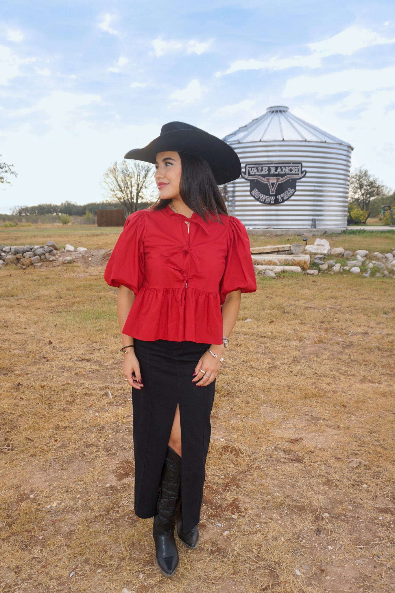 A woman posing in a field with a silo in the background modeling our scarlet red blouse that ties down the middle with bows and has bubble sleeves. She is wearing the top with a black denim long skirt with a front slit, a black cowboy hat and boots. 