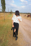 Full length photo of a woman modeling this cream colored v-neck blouse that ties with little black bows and stripes. It has bubble sleeves and ruffles at the shoulders. She is wearing it with our black denim skirt and black cowboy boots and is holding a wine bottle and wine glass.