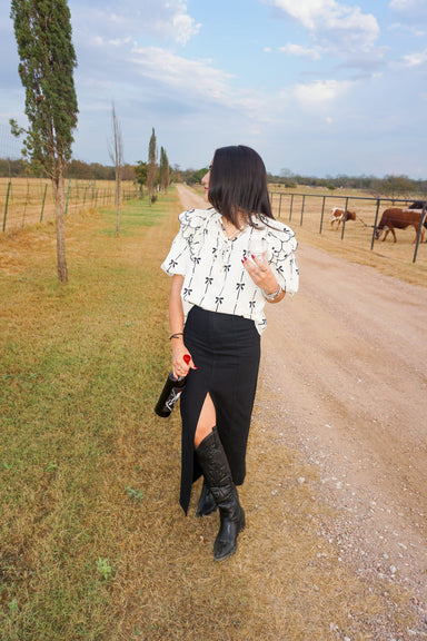 Full length photo of a woman modeling this cream colored v-neck blouse that ties with little black bows and stripes. It has bubble sleeves and ruffles at the shoulders. She is wearing it with our black denim skirt and black cowboy boots and is holding a wine bottle and wine glass.