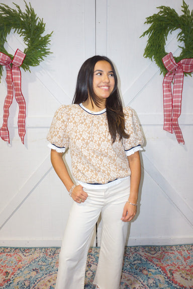 A woman modeling a beige top with a white rose pattern all over. The neckline, sleeves and waist all have a navy and white trim.