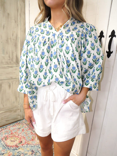 Up close photo of our white 3/4 sleeve, v-neck blouse that has light blue flowers with green leaves all over and our white linen shorts.