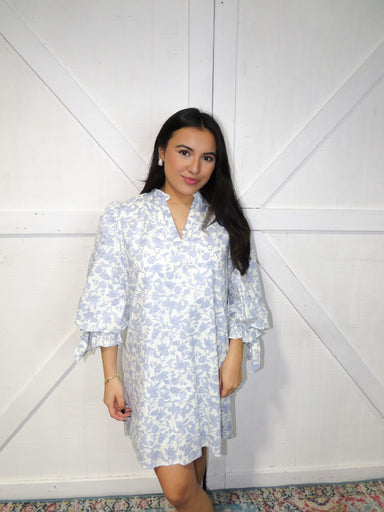 Photo of our model wearing our off-white, 3/4 sleeve, v-neck short (just above the knees) dress that has bows at the sleeves and a light blue vine pattern all over.