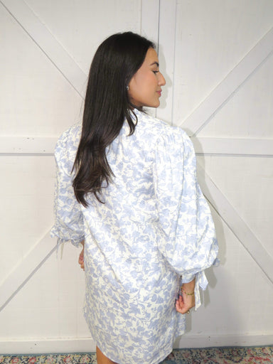 Photo of our the back of our off-white, 3/4 sleeve, v-neck short (just above the knees) dress that has bows at the sleeves and a light blue vine pattern all over.